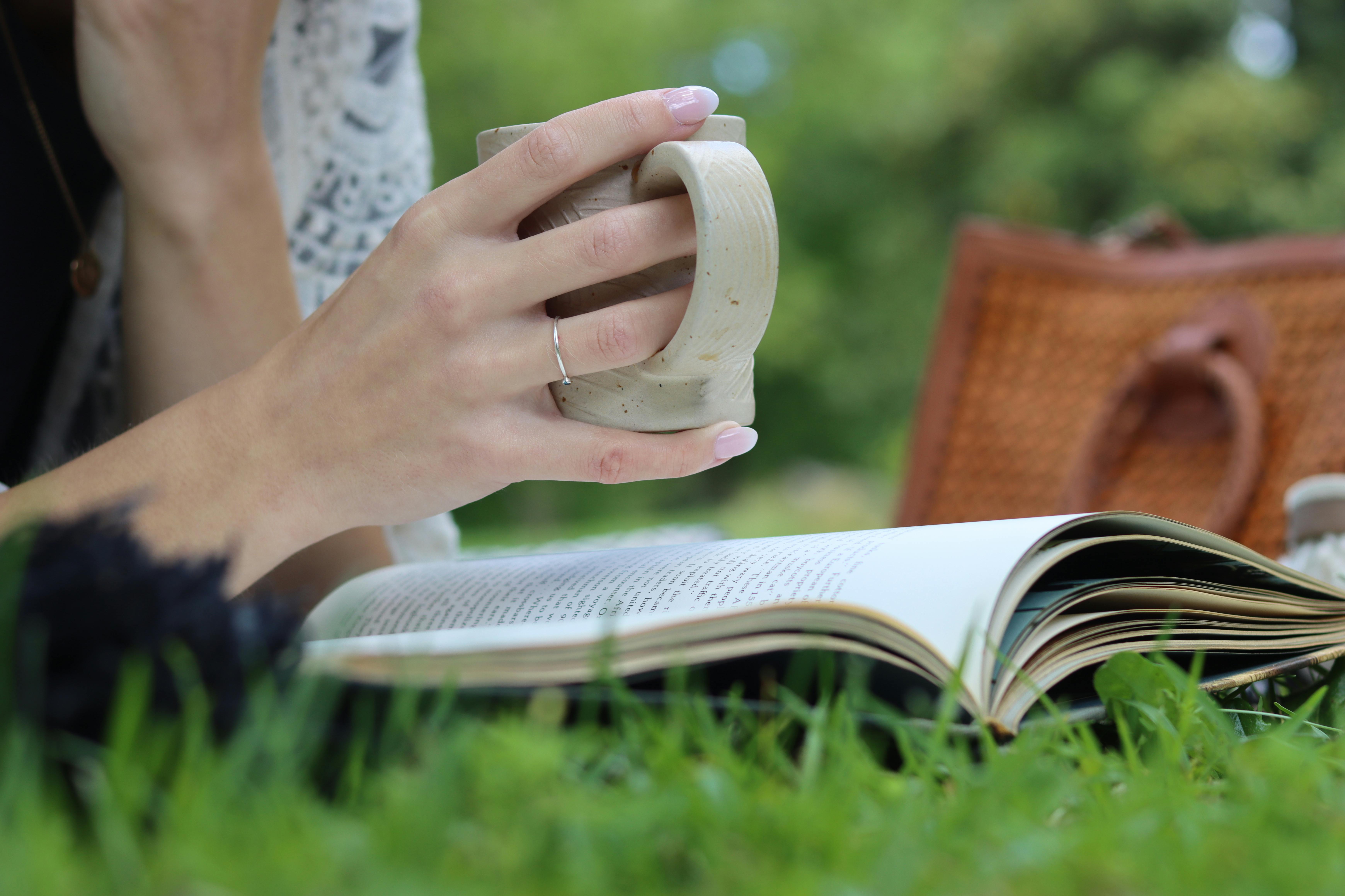 Tangan Memegang Cangkir Sambil Membaca Buku Di Taman
